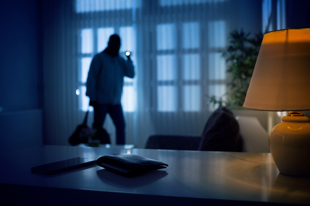 man in dark home with curtains drawn, holding a flashlight pointing at a wallet and phone on a table in the foreground